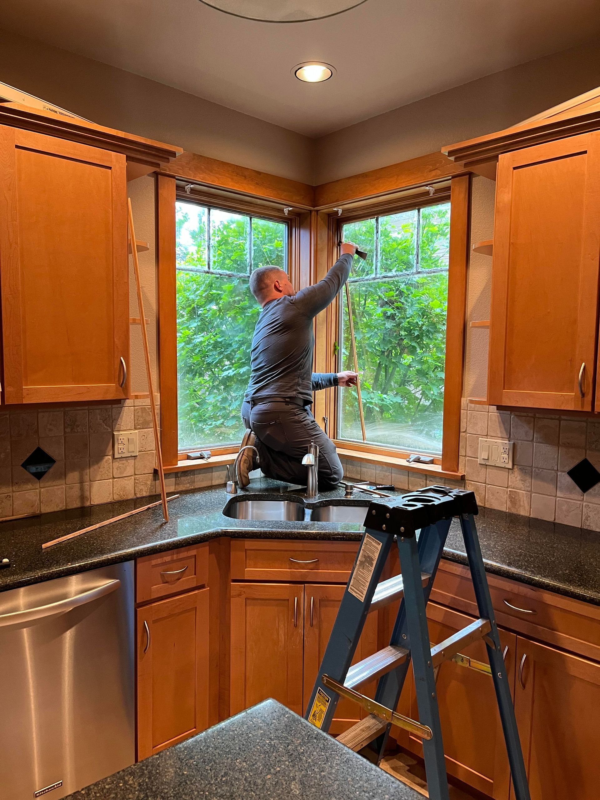 A man is standing on a ladder in a kitchen cleaning a window.