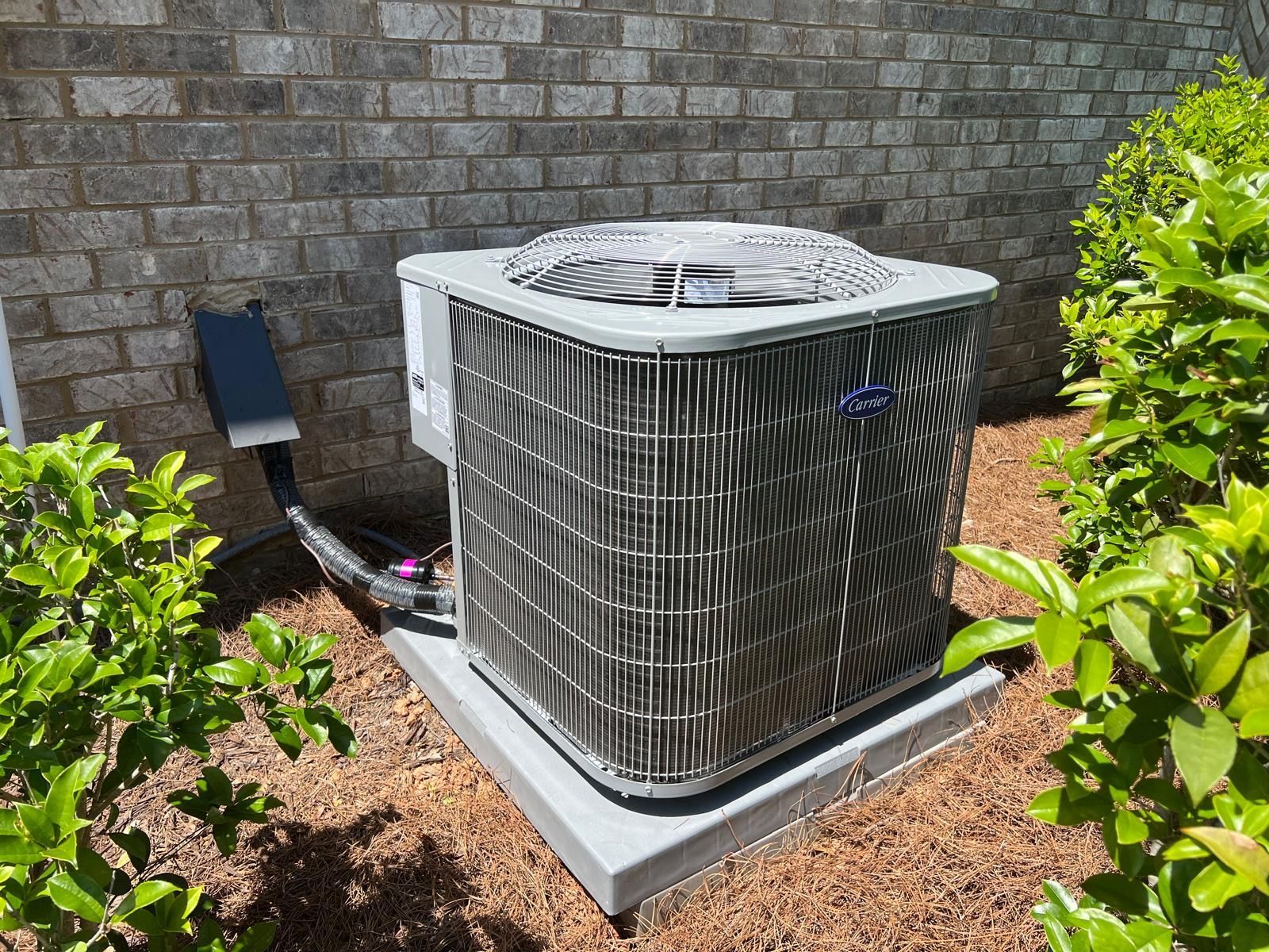 Outdoor HVAC condenser unit beside a brick wall, surrounded by mulch and shrubs.