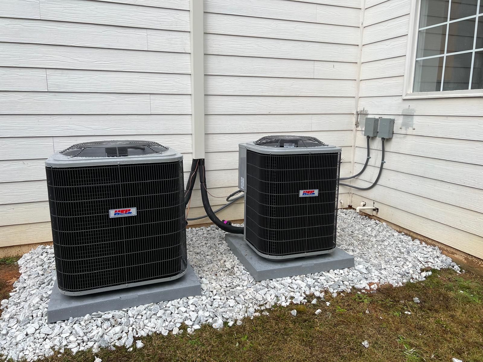 Two black outdoor HVAC units on concrete pads beside a white house wall
