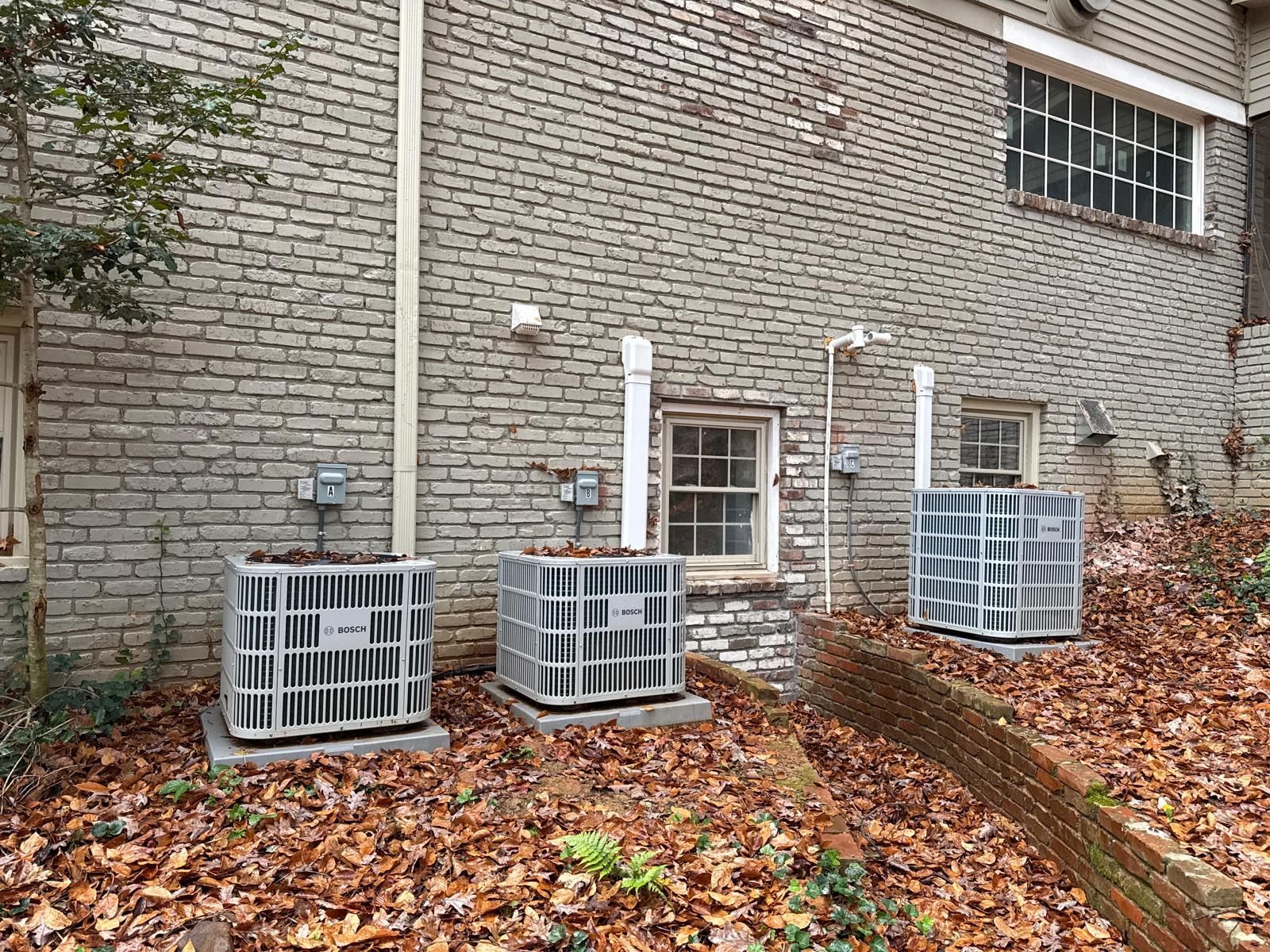Three outdoor HVAC units beside a brick building with leaf-covered ground and a sloped walkway