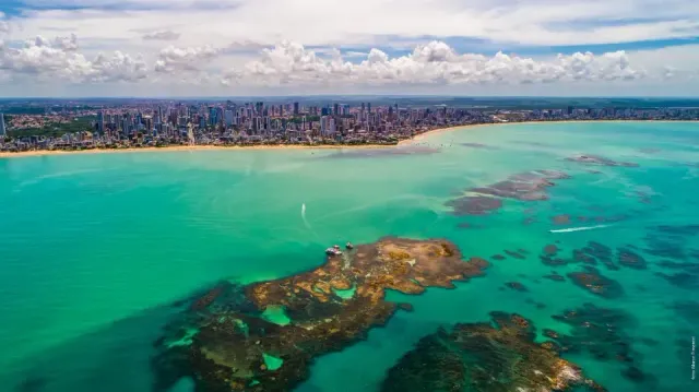 Vista aérea do oceano turquesa, recifes de coral e horizonte de uma cidade costeira sob um céu nublado.