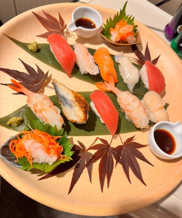 Sushi platter with various nigiri, soy sauce, and garnish on a wooden plate.