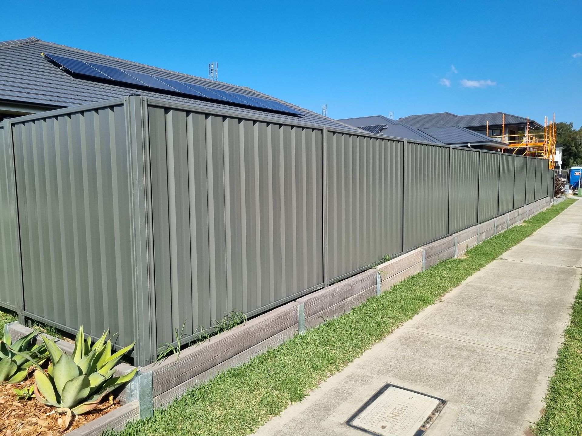 Green corrugated metal fence along a sidewalk, with grass and residential homes visible.