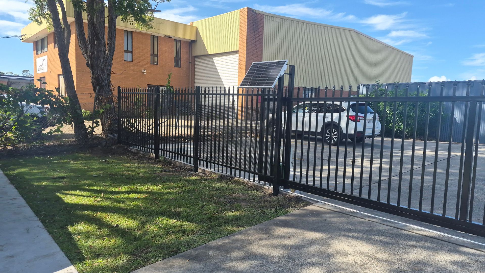 Black metal fence beside a sidewalk and parking lot in front of a tan building with white canopy — Strike Fencing & Gates In Davistown, NSW