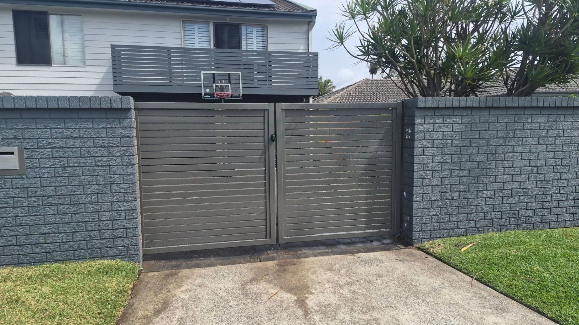 Gray metal driveway gate in front of a house, with a basketball hoop above it and brick walls on both sides — Strike Fencing & Gates In Terrigal, NSW