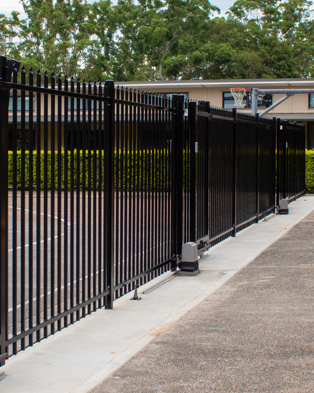 Black metal fence alongside a concrete path, extending towards a building with green foliage visible behind it. — Strike Fencing & Gates In Tuggerah, NSW