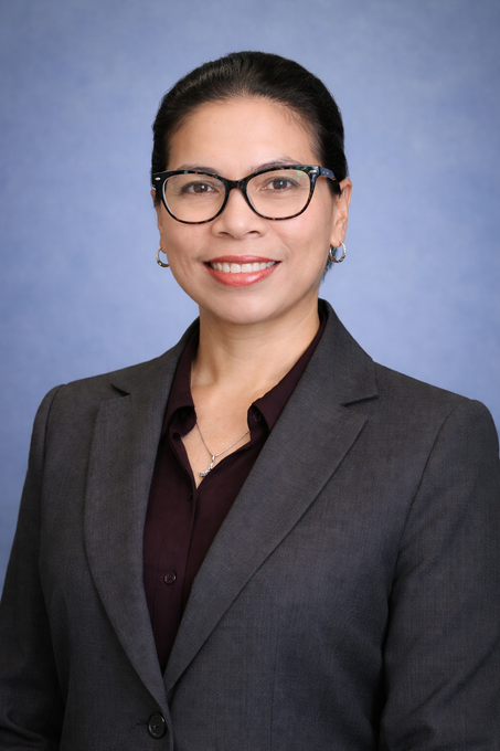 Professional portrait of a woman in glasses and gray blazer, smiling against a blue studio backdrop