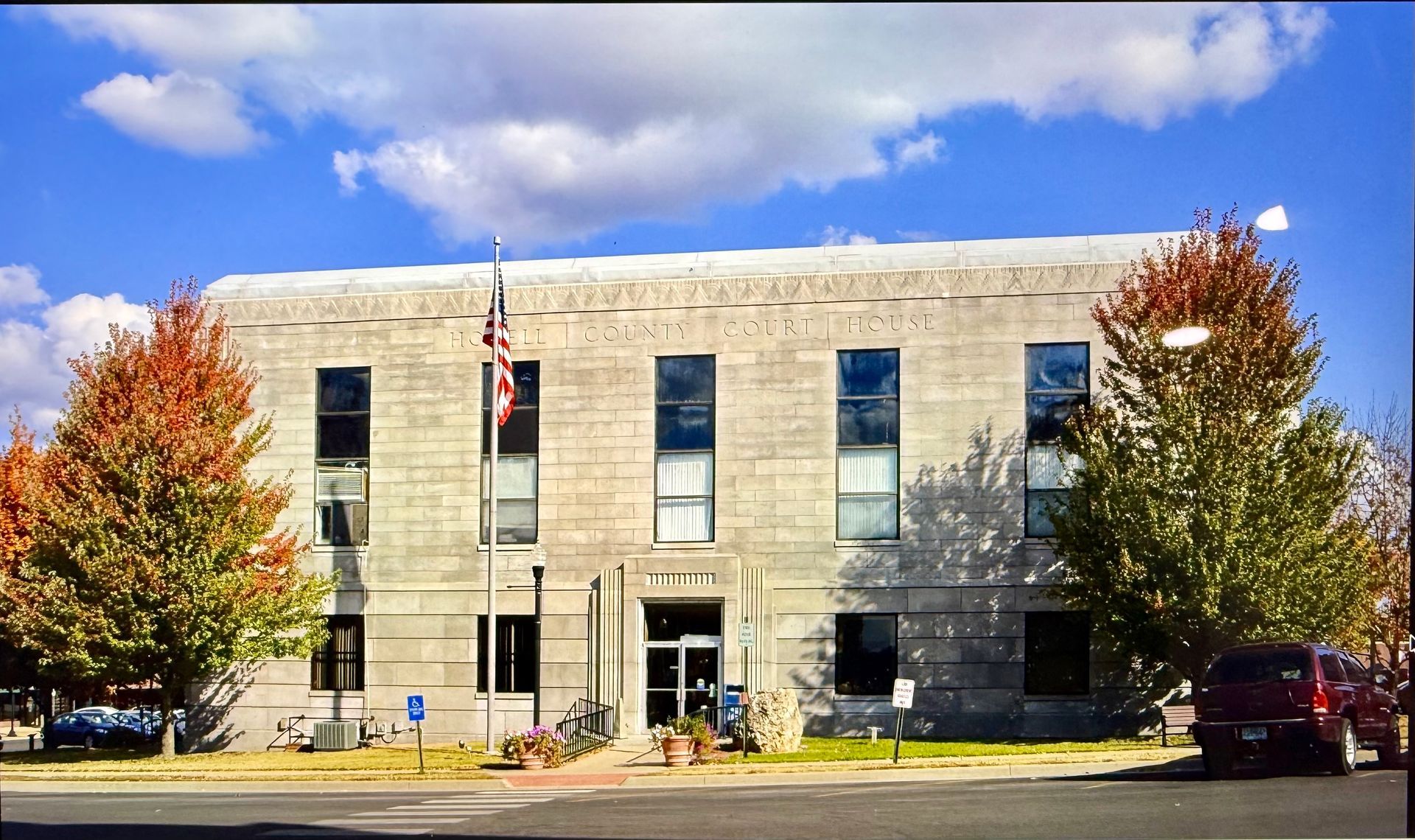 Stone civic building with an American flag, tall windows, and autumn trees in front