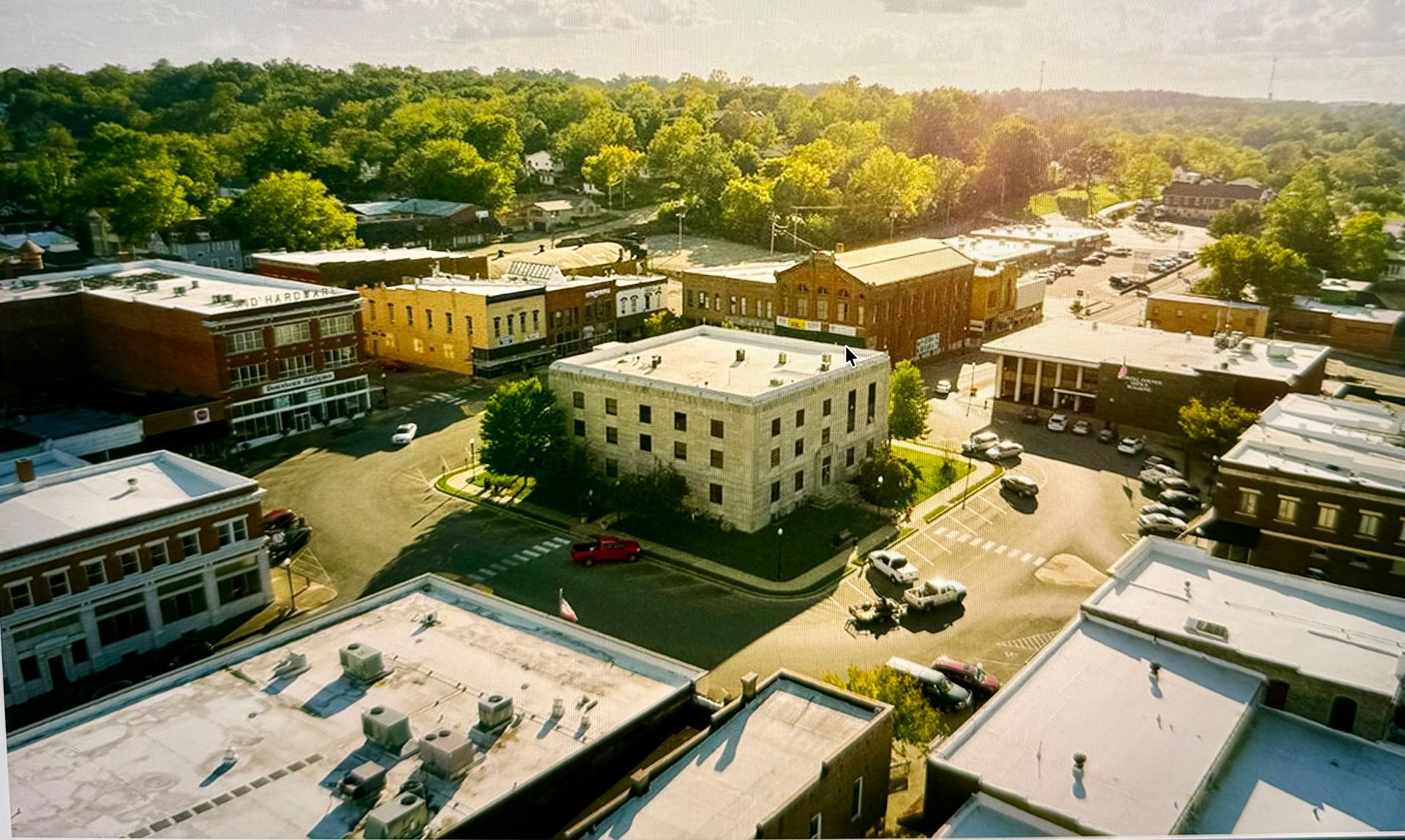 Aerial view of a small town center with brick buildings, snowy rooftops, and trees in warm sunlight