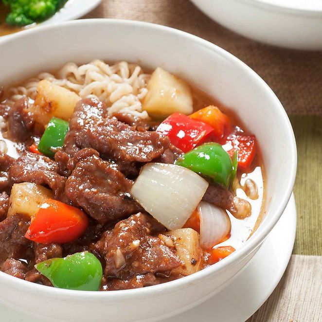 Bowl of beef and vegetables over noodles, with red, green, and white bell peppers, on a table