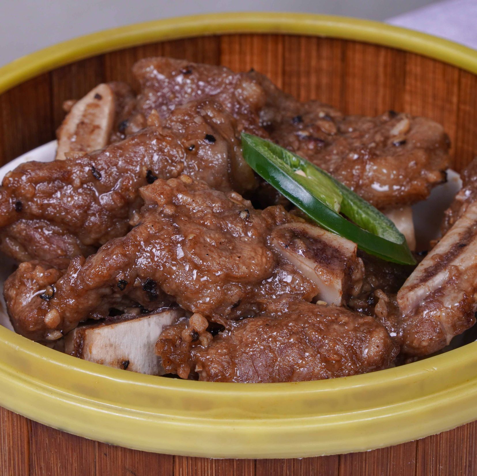 Braised meat ribs in a yellow-rimmed bowl, topped with sliced green chili peppers.