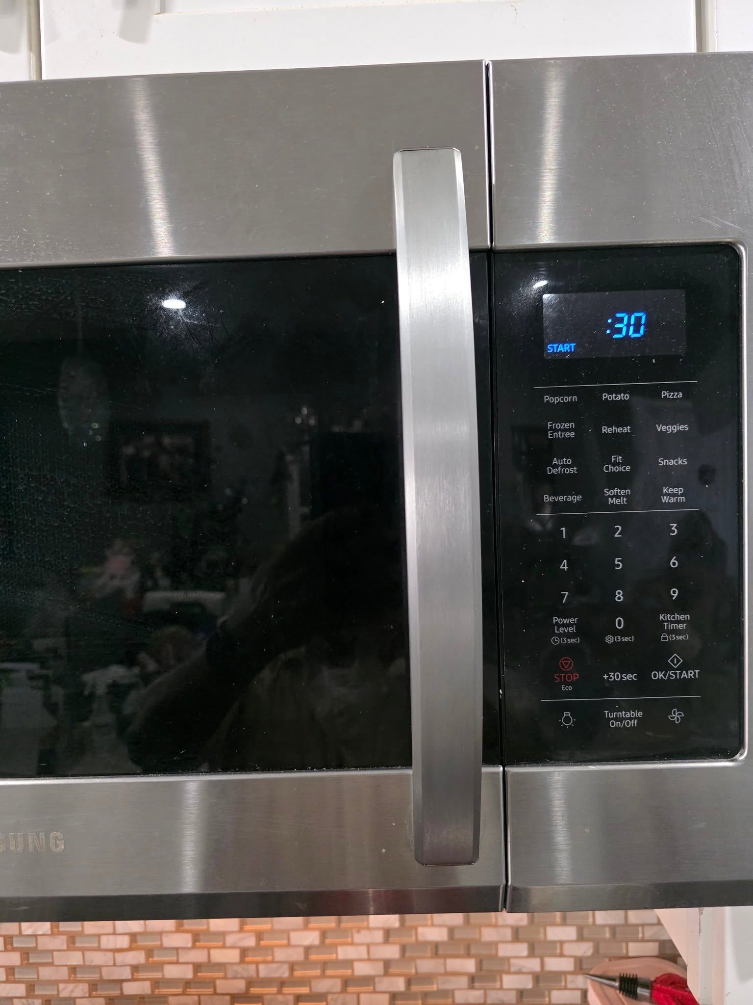 Stainless steel microwave with black door and digital control panel above a tiled backsplash.
