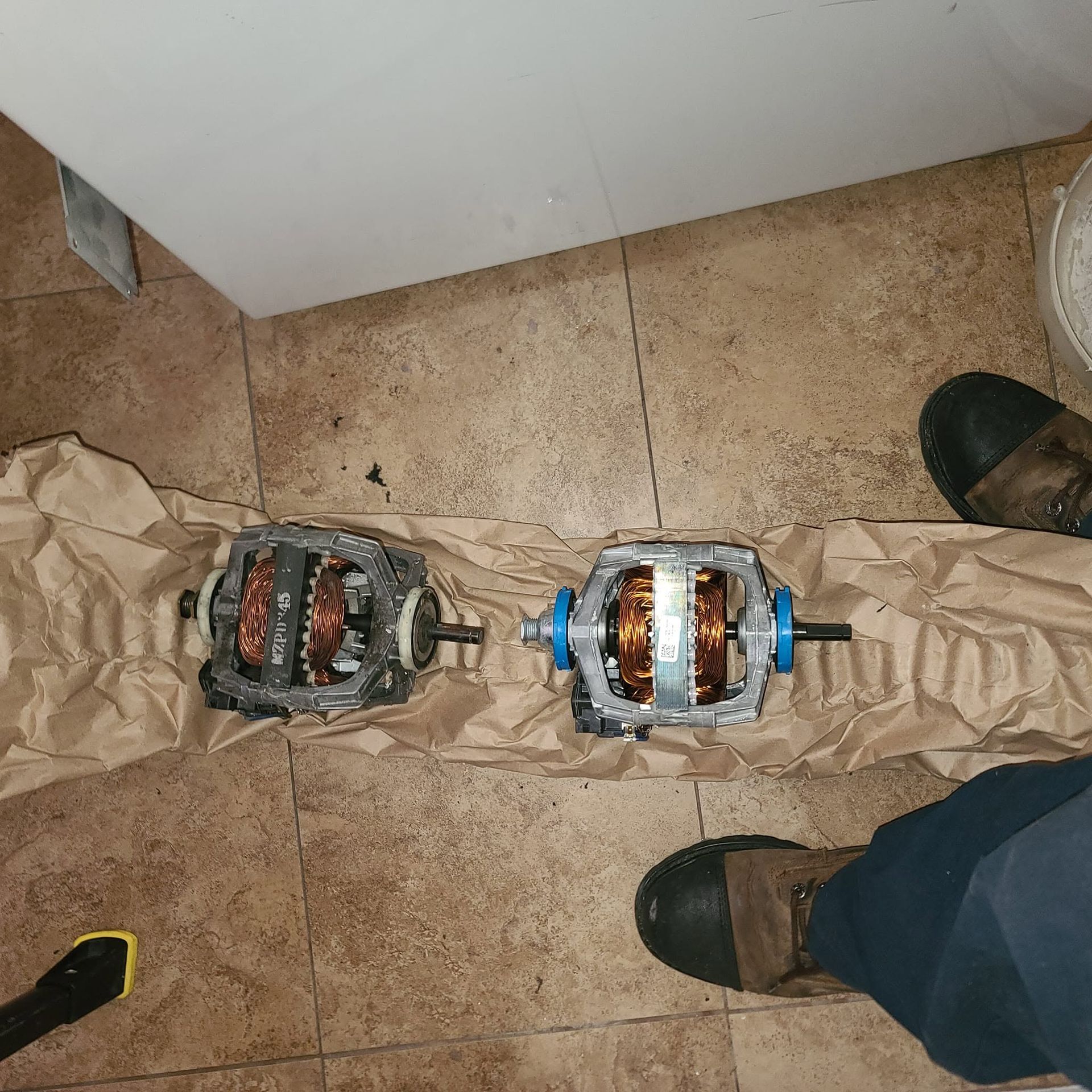 Two electric motor rotors on cardboard on a tiled floor, with shoes visible nearby