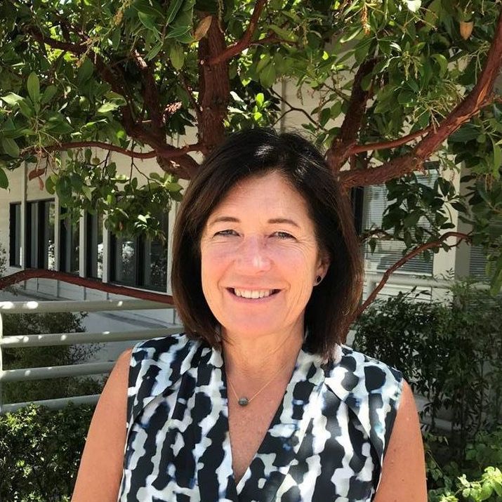 Woman with brown hair smiles in a black and white patterned top, outdoors near leafy tree and building.