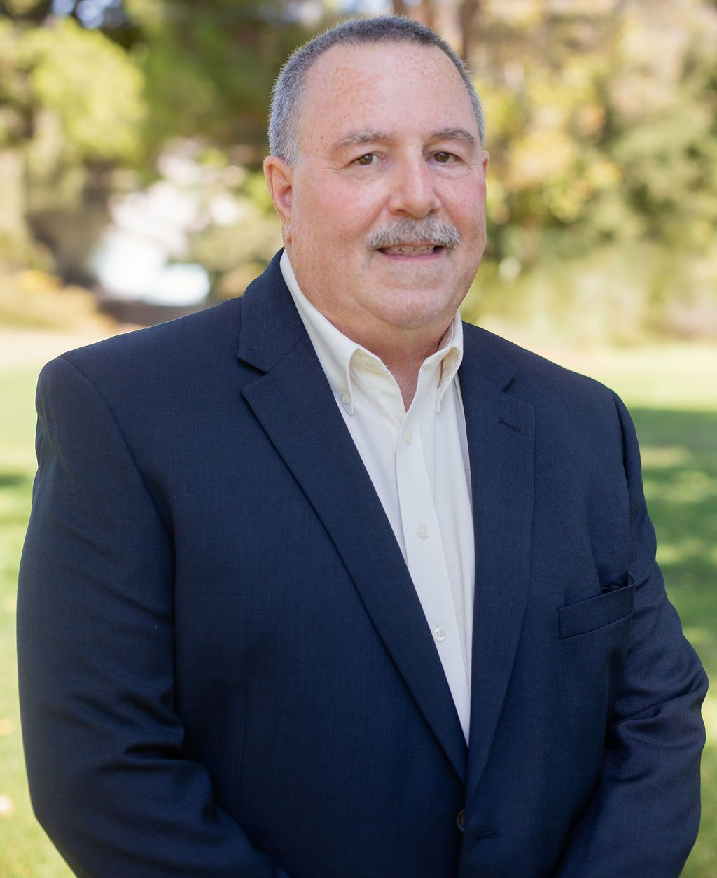 Man in a navy blazer and open-collared cream shirt, smiling outdoors in front of trees and a pond.