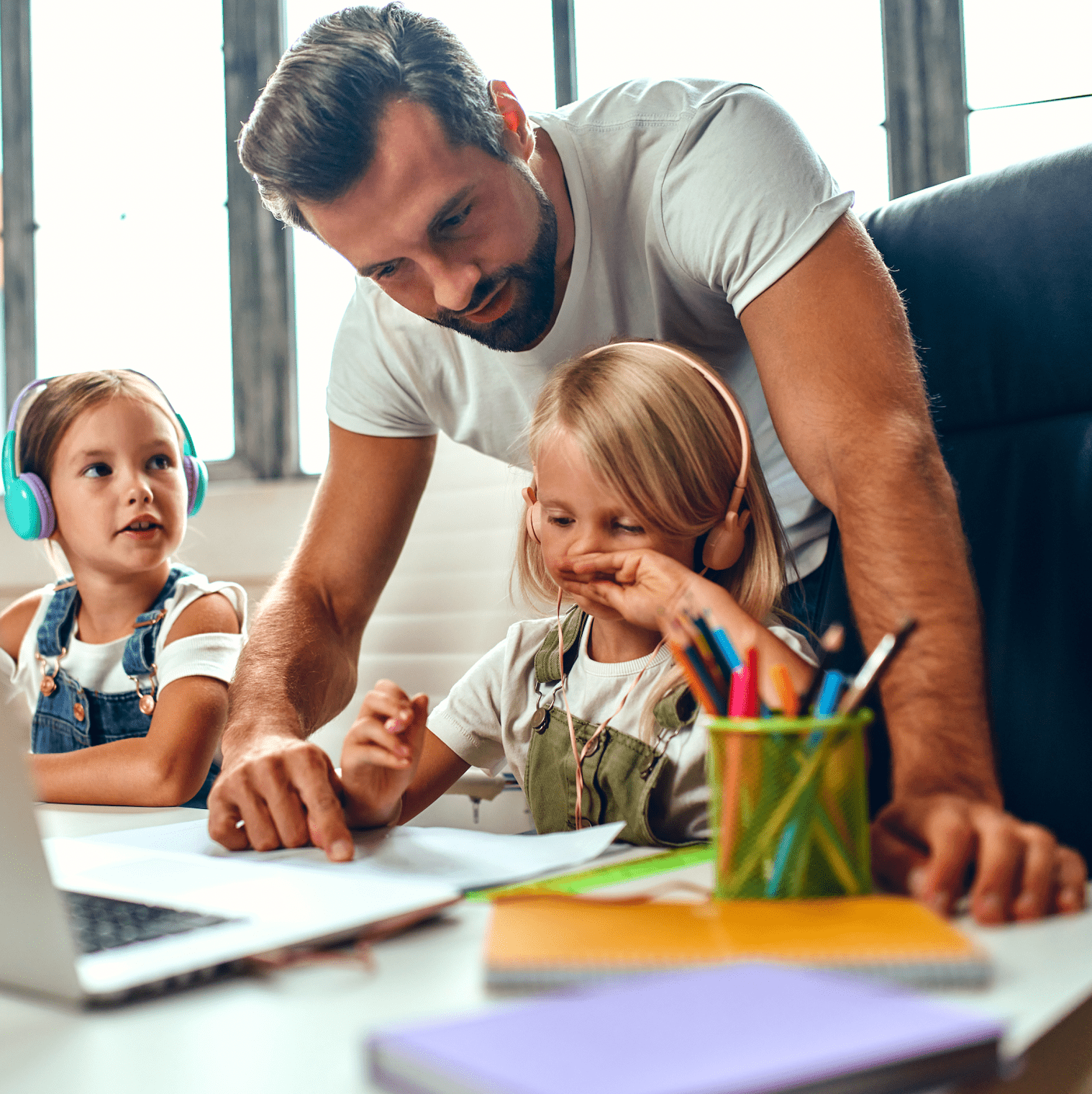 A man helps two young children with schoolwork at a desk. One child wears headphones.