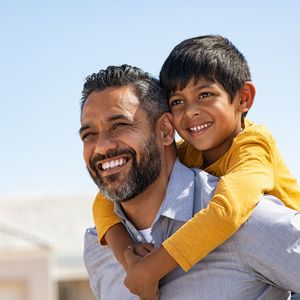 Man smiling, carrying a child on his back outdoors, blue sky.