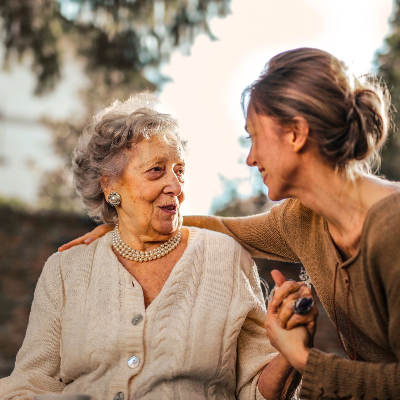 Woman comforts an elderly woman outdoors, hand on shoulder, looking at each other, warm tones.