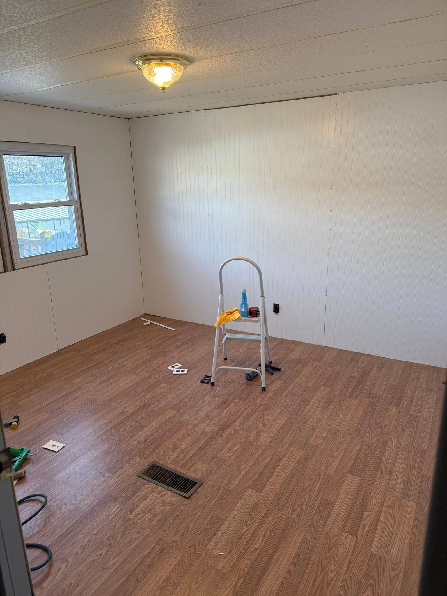 Empty room with wood-pattern floor, white walls, window, ceiling light, and a step ladder in the center