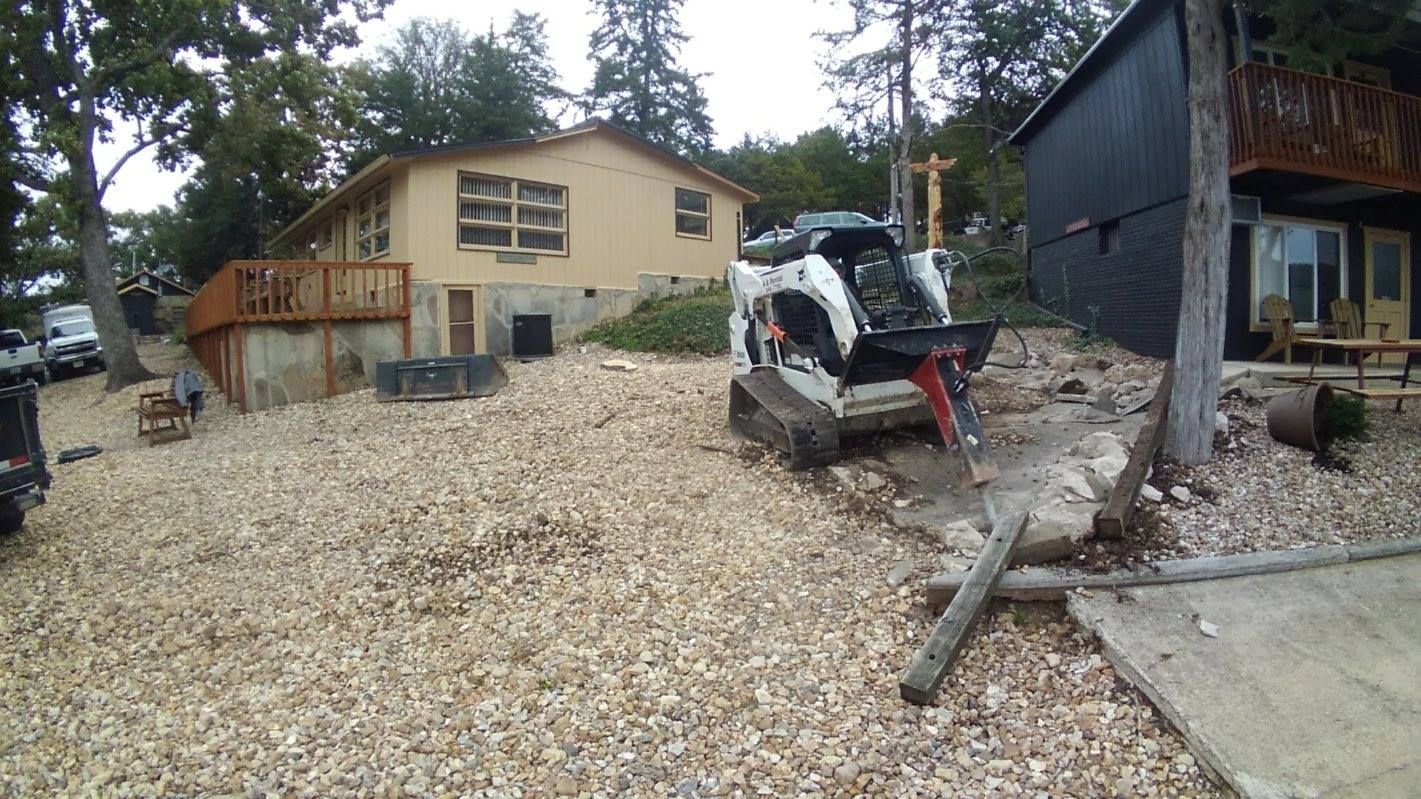A Bobcat skid-steer loader removing rubble in front of a house next to a dark cabin; outdoor scene.