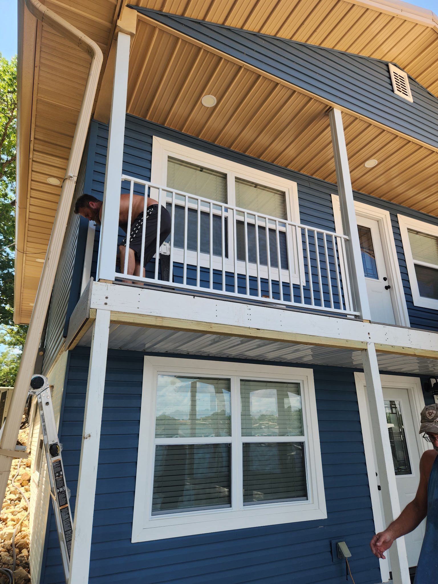 Person on a ladder, working on the balcony of a blue two-story house with white trim.