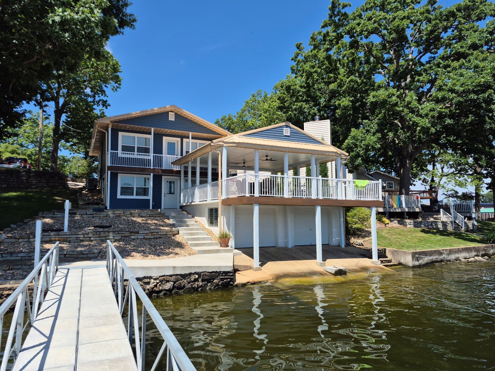 Two-story blue house with porch overlooking a lake, attached dock in the water, sunny day.