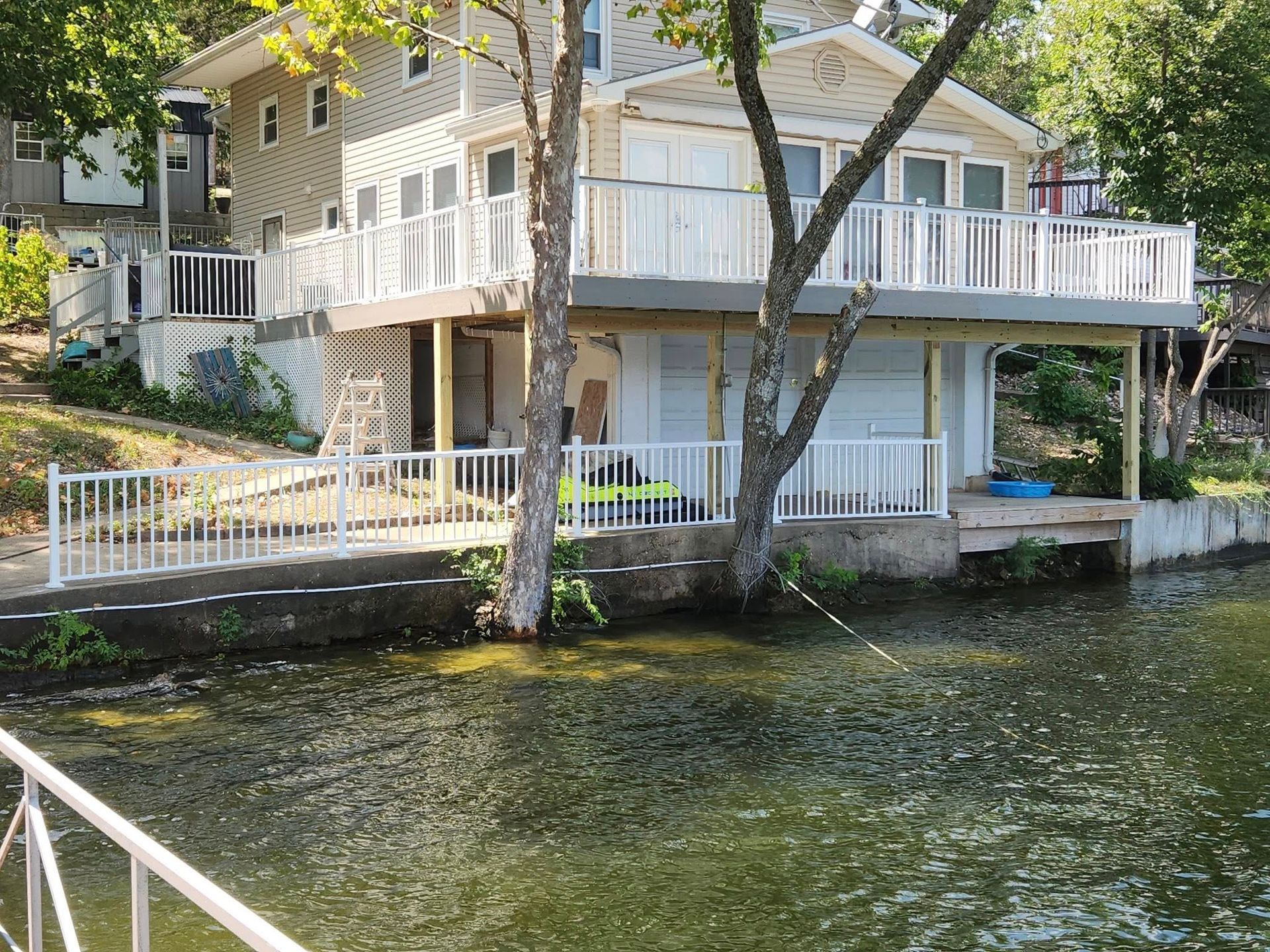 Lakeside house with white railings and two-story deck, partially obscured by trees; water in the foreground.