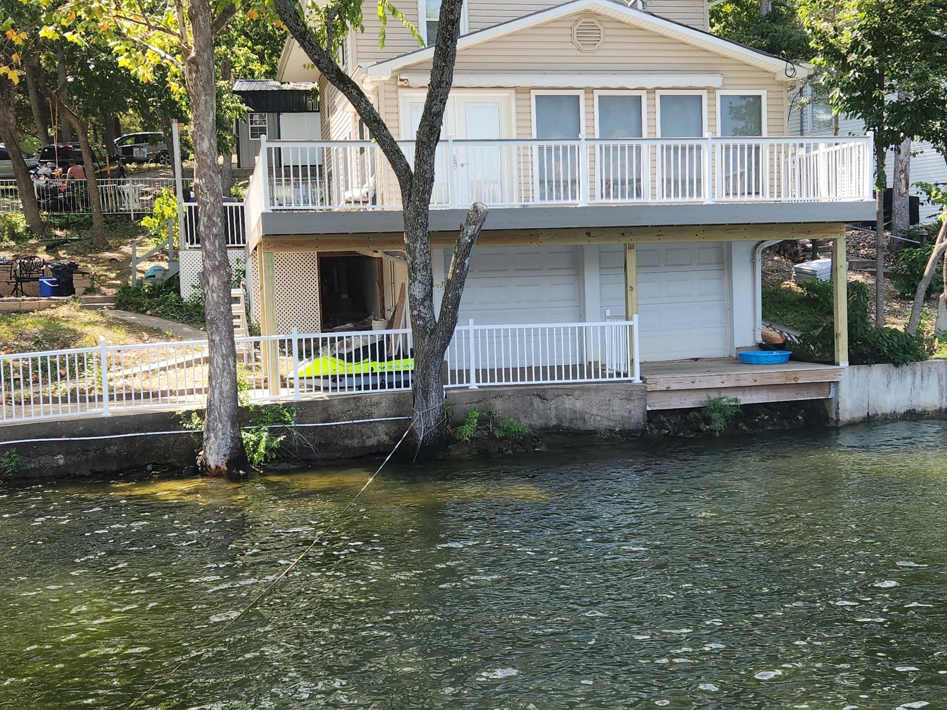 Two-story waterfront house with white railings and garage, viewed from the water. Green trees surround the house.