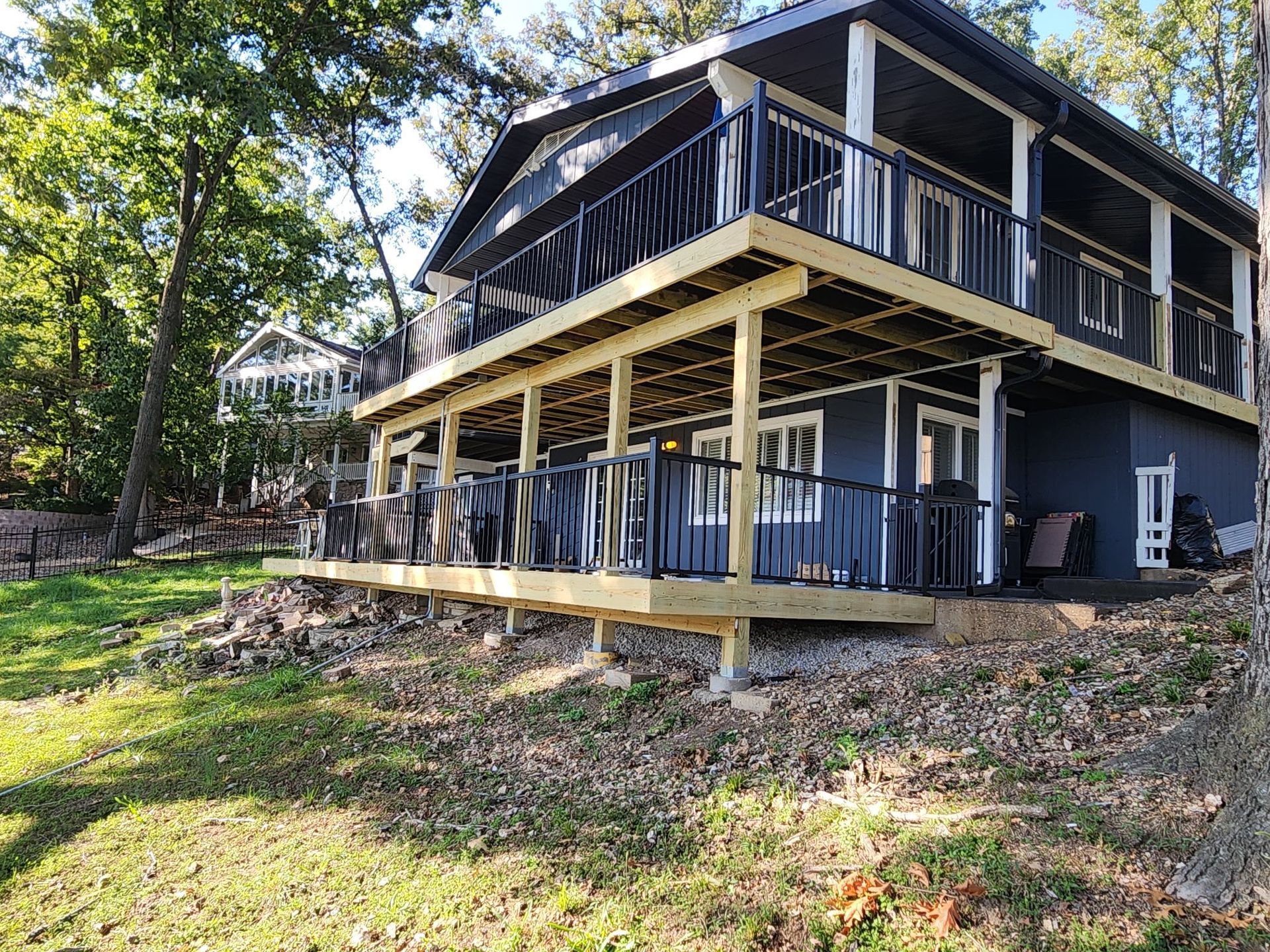Two-story house with black trim, two decks, and a gray exterior. Green lawn, trees, and sky in the background.