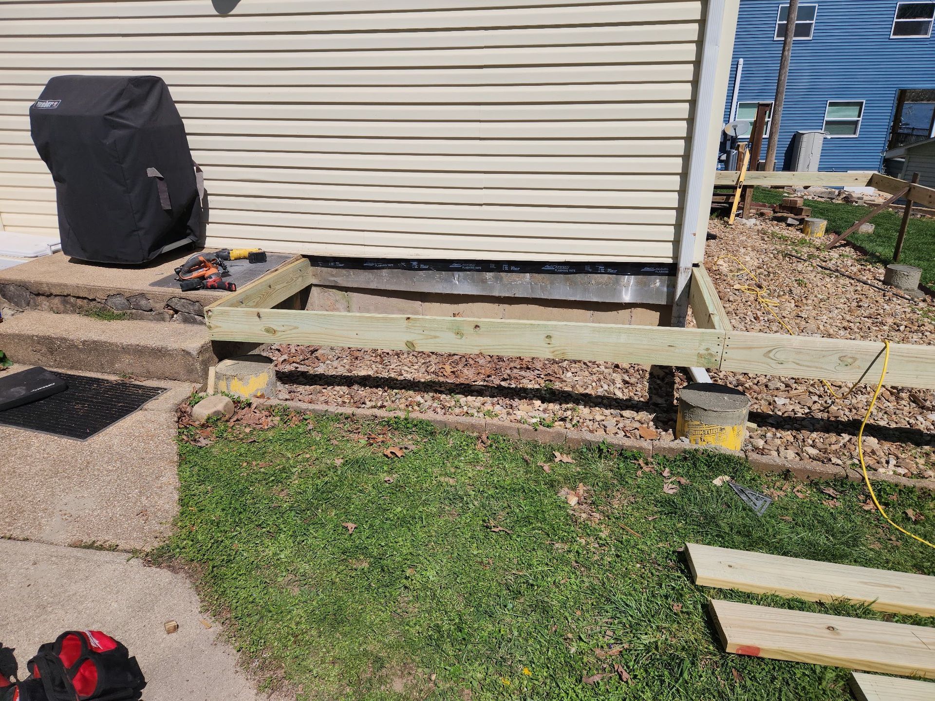 Backyard deck with a wooden bench, beige house siding, grass, gravel, and lumber on the ground