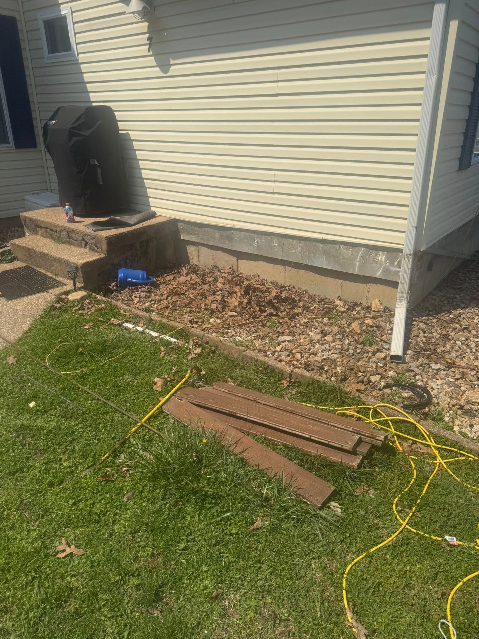 Backyard beside house siding with yellow extension cord and brown wooden planks on grass and rocks.