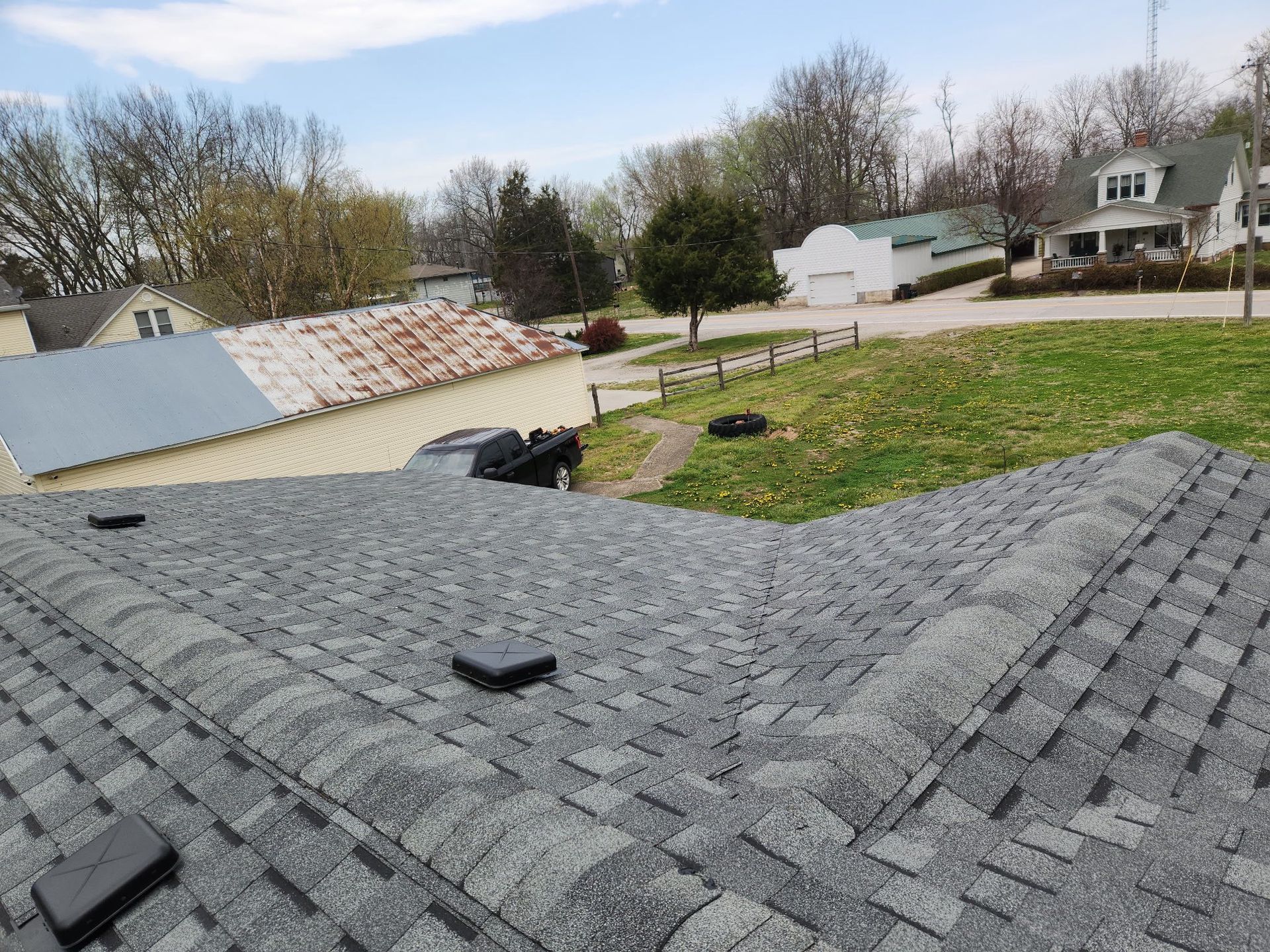 Rooftop with gray shingles overlooking a grassy yard, fences, trees, and nearby houses under a cloudy sky