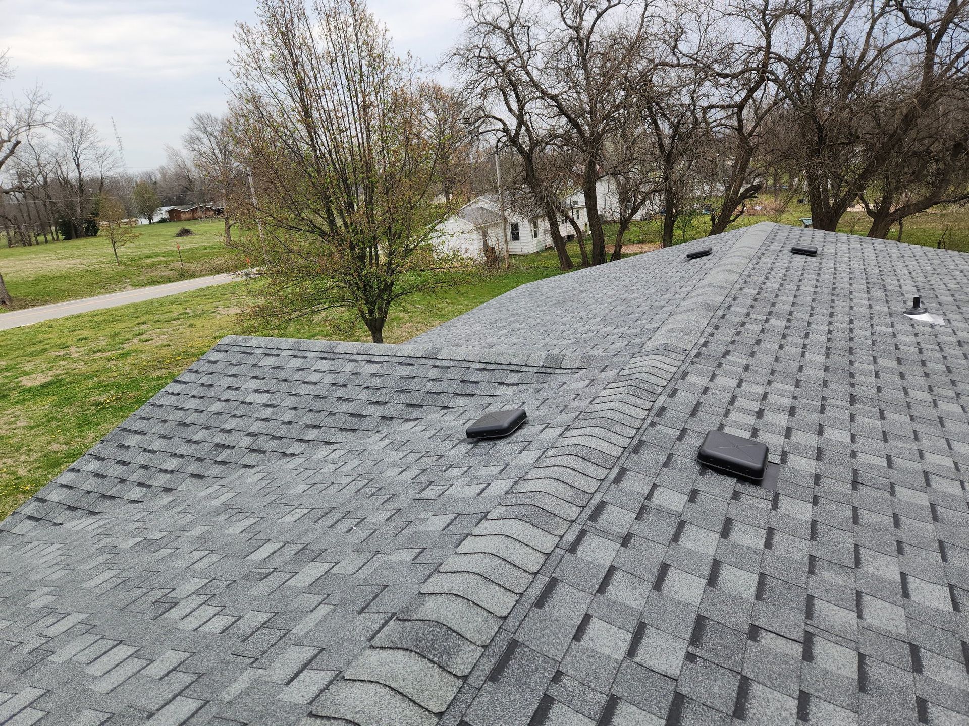 Gray shingled roof with two vents beside a grassy yard and leafless trees