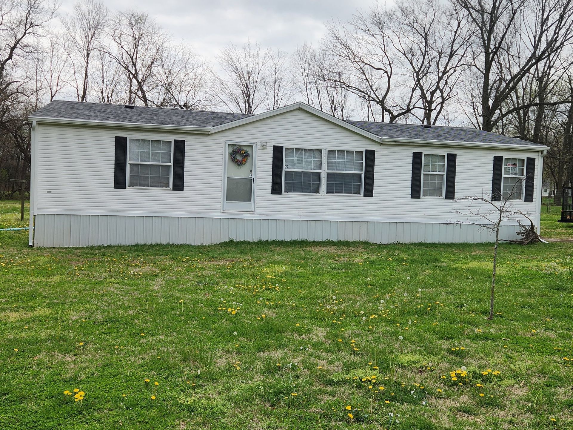 White mobile home with black shutters in a grassy yard under bare trees