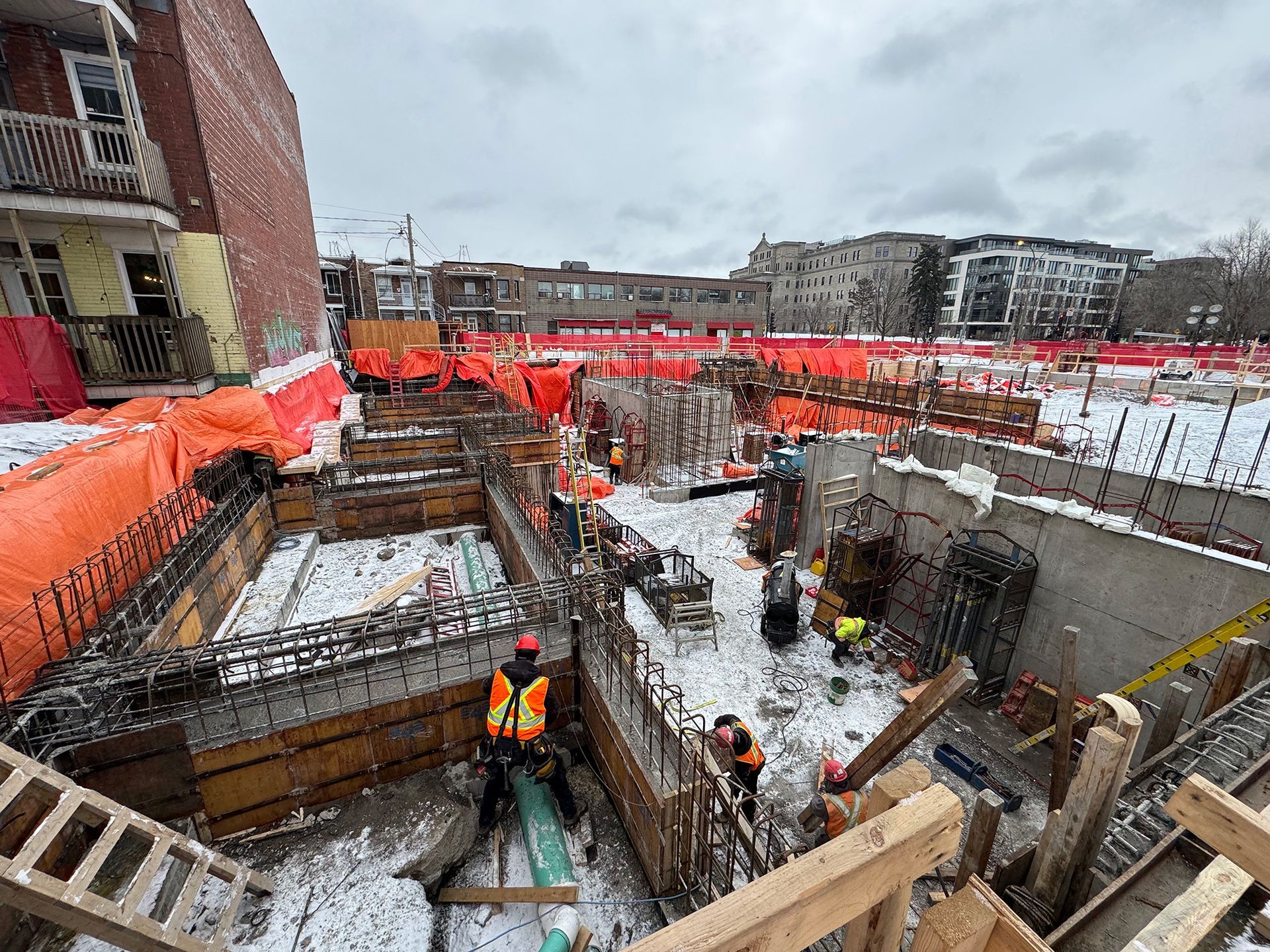 Chantier de construction avec ouvriers, barrières orange et coffrages en béton, probablement pour les fondations d'un bâtiment, en hiver.