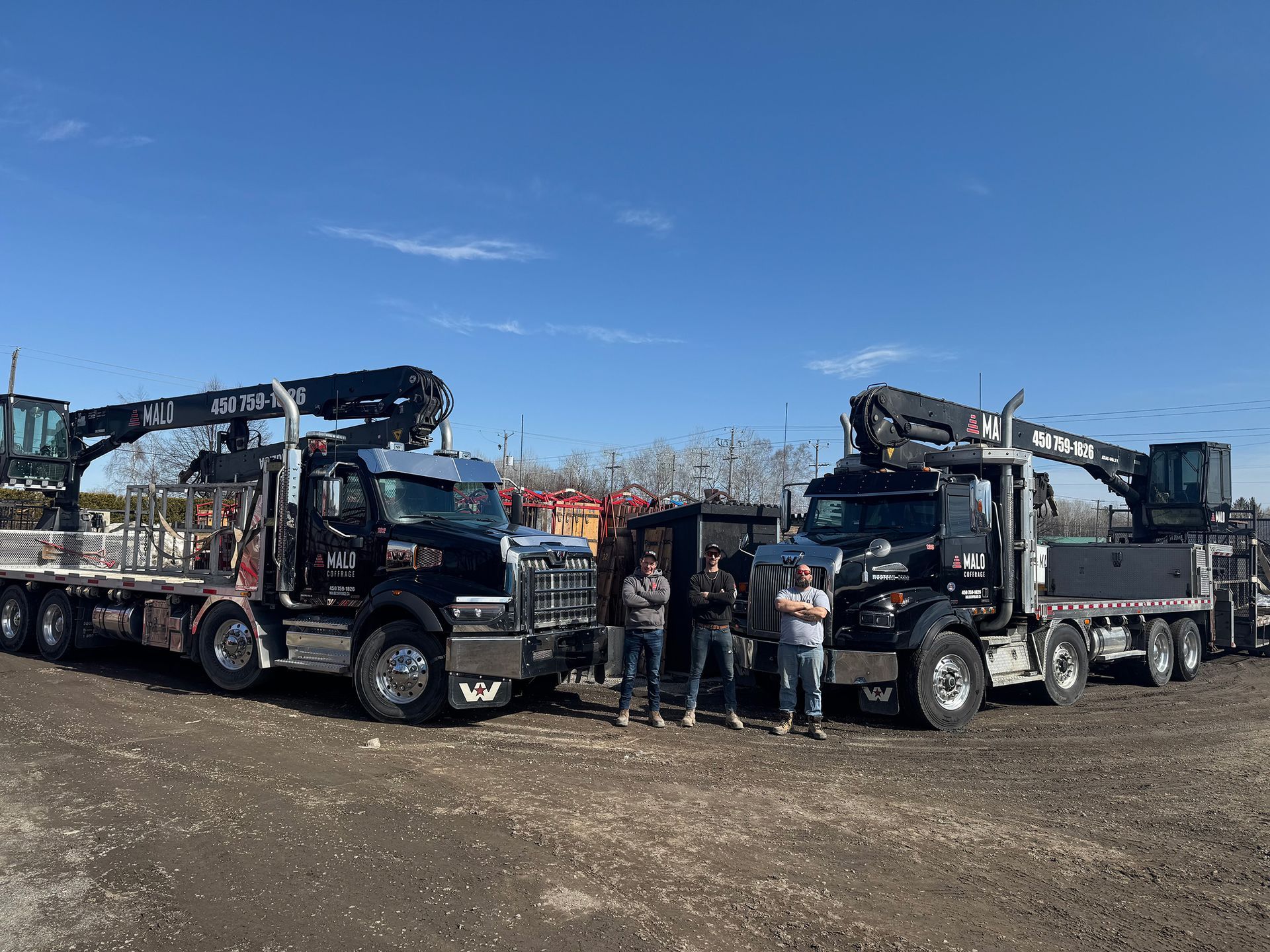 Deux camions-grues noirs à plateau ; trois hommes debout devant eux sous un ciel bleu.