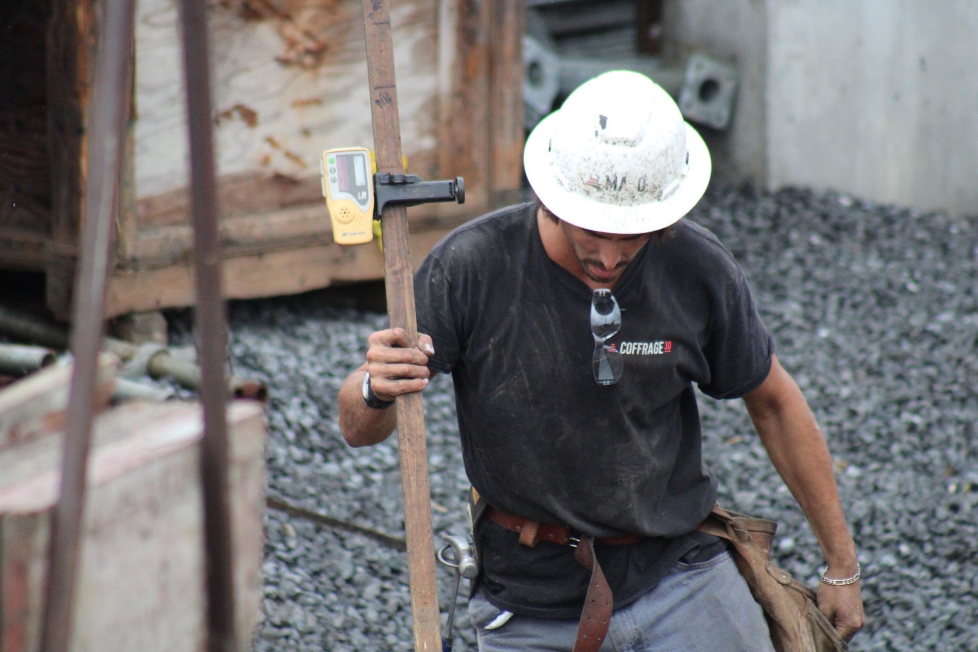 Un ouvrier du bâtiment, portant un casque blanc et une chemise noire, transporte un niveau à bulle sur un chantier.