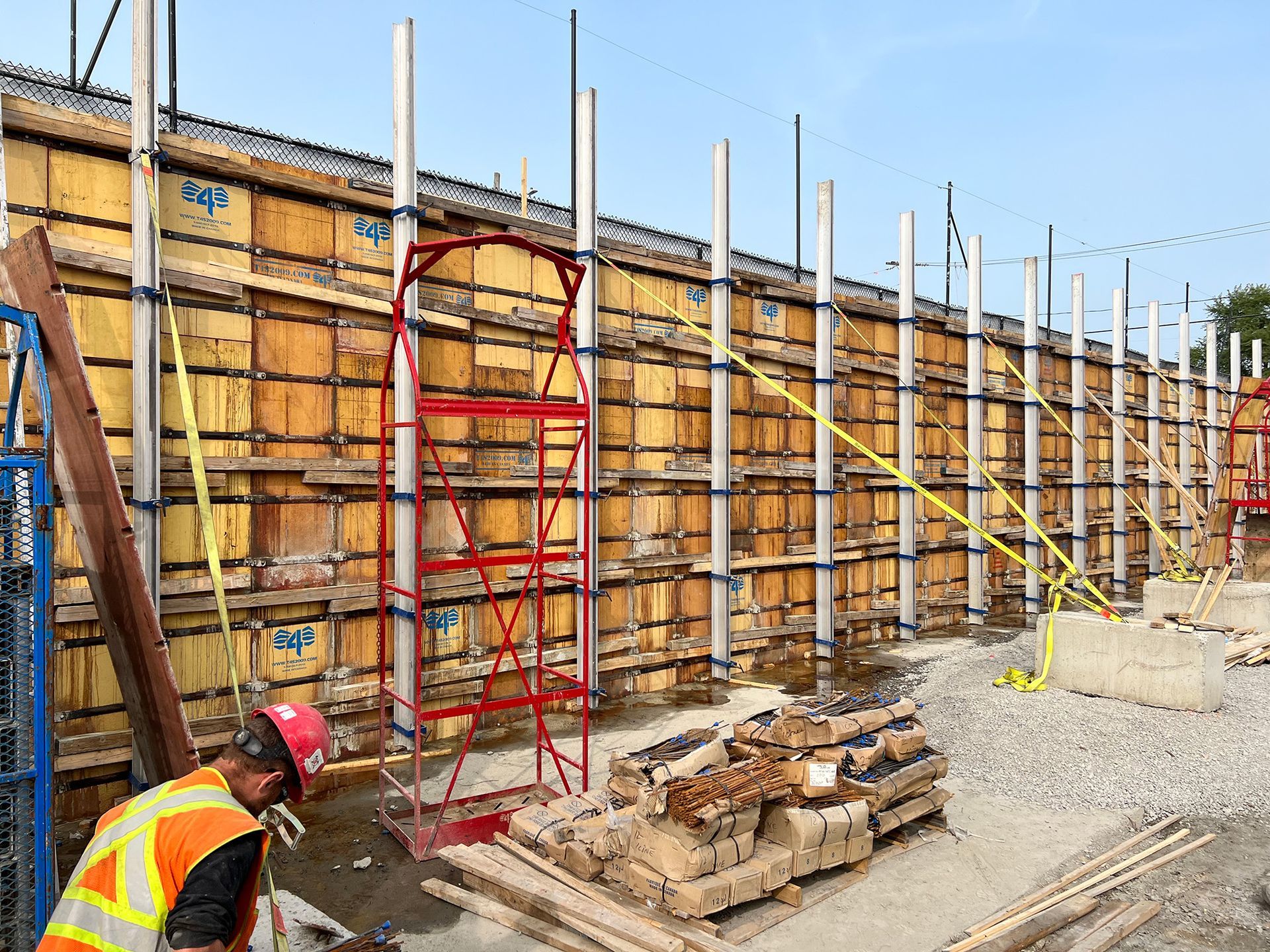 Chantier de construction avec un ouvrier en tenue de sécurité près d'un coffrage et d'un échafaudage en bois.