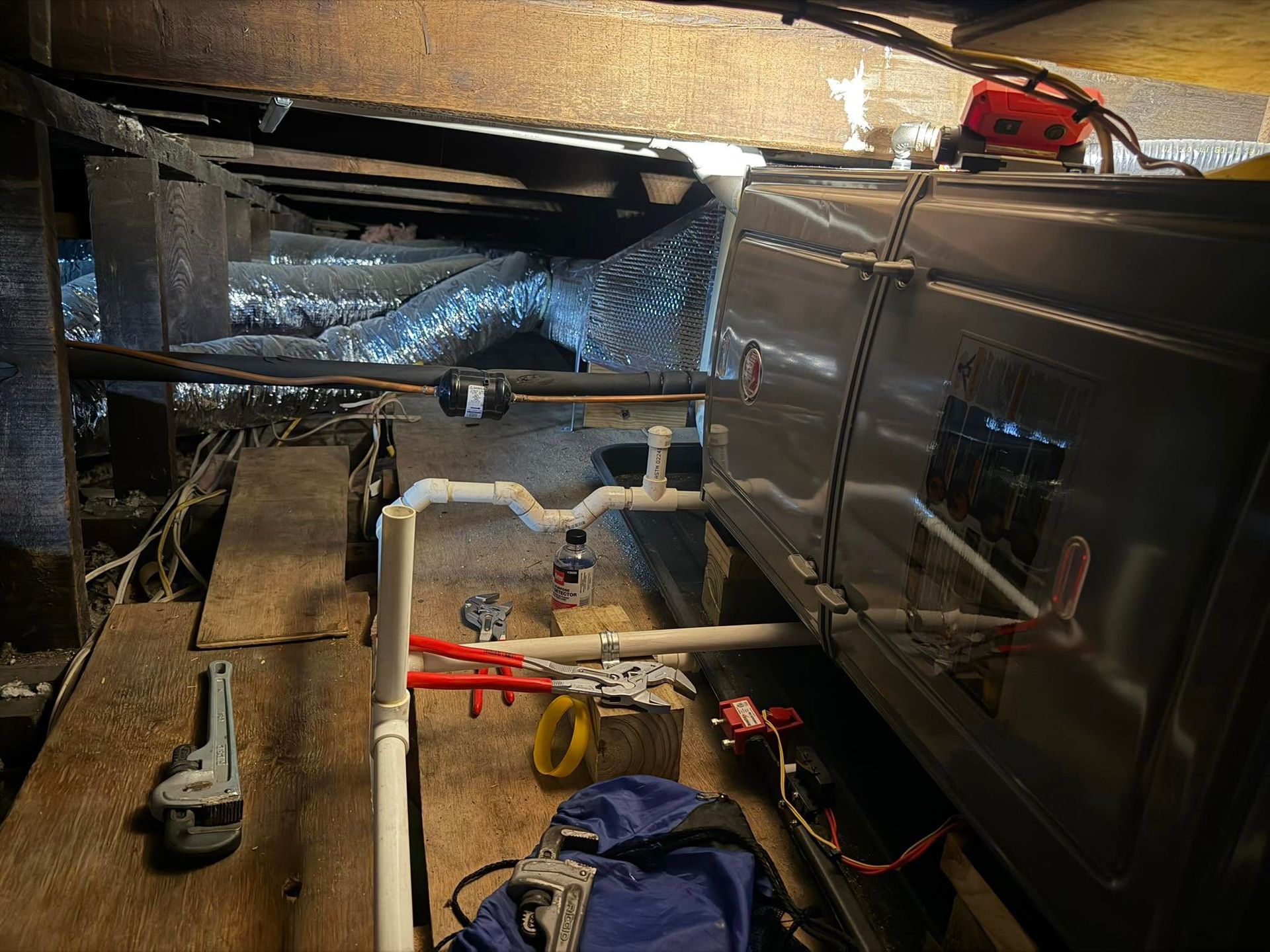 Plumbing beneath a floor: pipes, valves, and tools in a dark crawlspace with ductwork above.