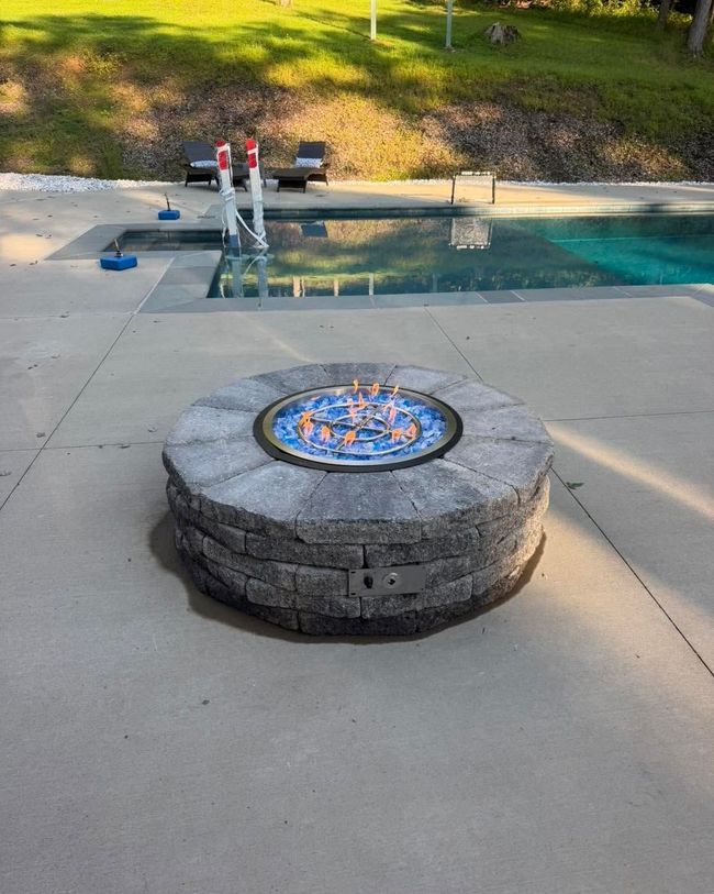 Round stone fire pit by a pool with a small blue flame, in a concrete backyard patio.