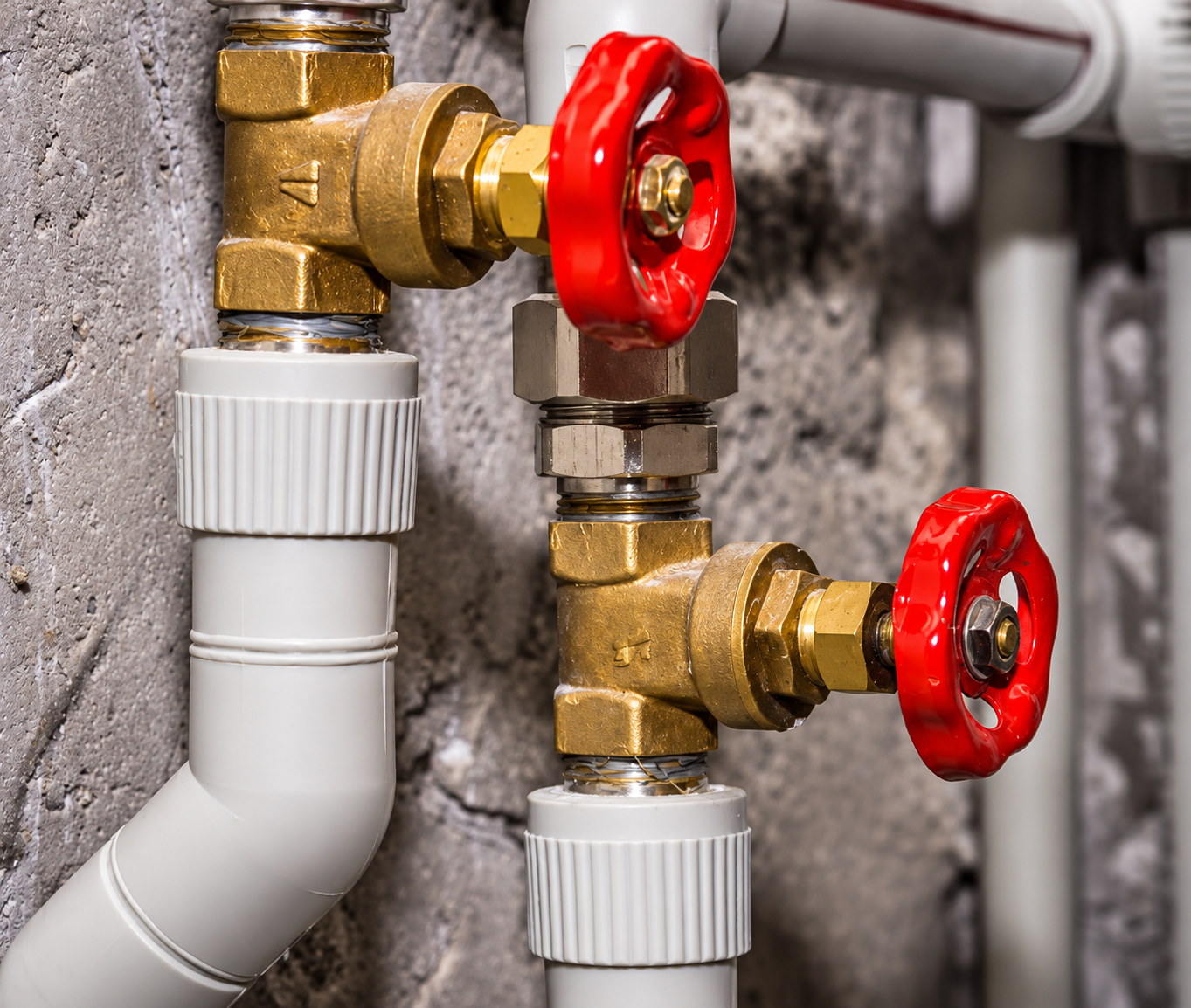 Close-up of brass water pipes with red shutoff valves and white PVC joints on a concrete wall