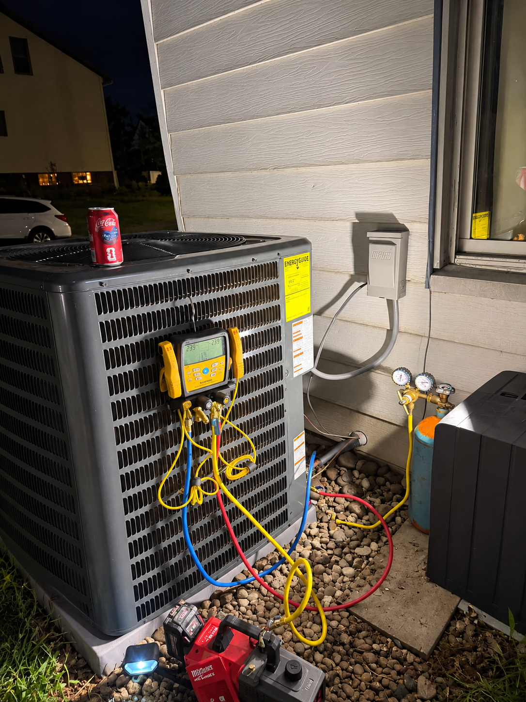 Outdoor air conditioner unit with gauges, hoses, and tools attached against a house wall at night