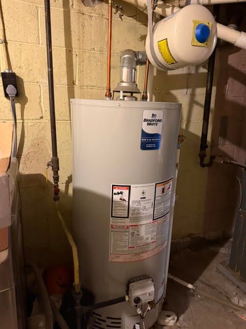 Water heater in a basement utility room with exposed pipes and a small expansion tank above it