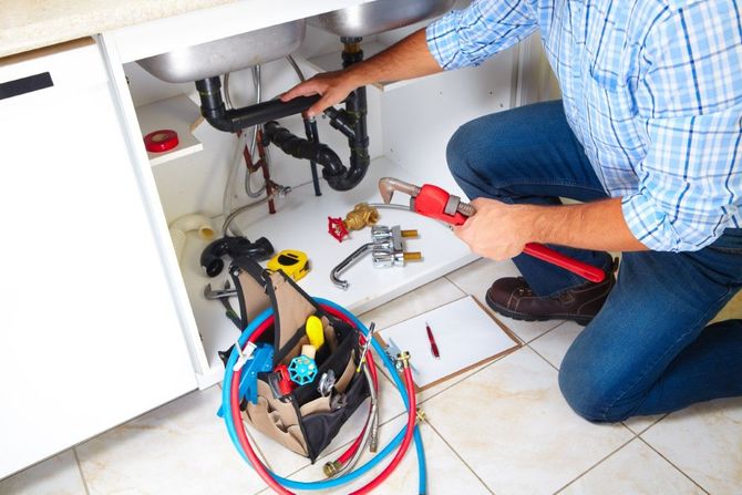 Plumber kneeling under a sink, using tools to repair pipes with colorful hoses and supplies on the floor