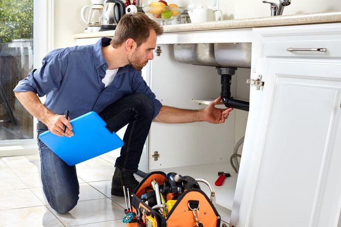 Plumber inspecting under-sink pipes in a white kitchen, holding a blue folder and tools nearby