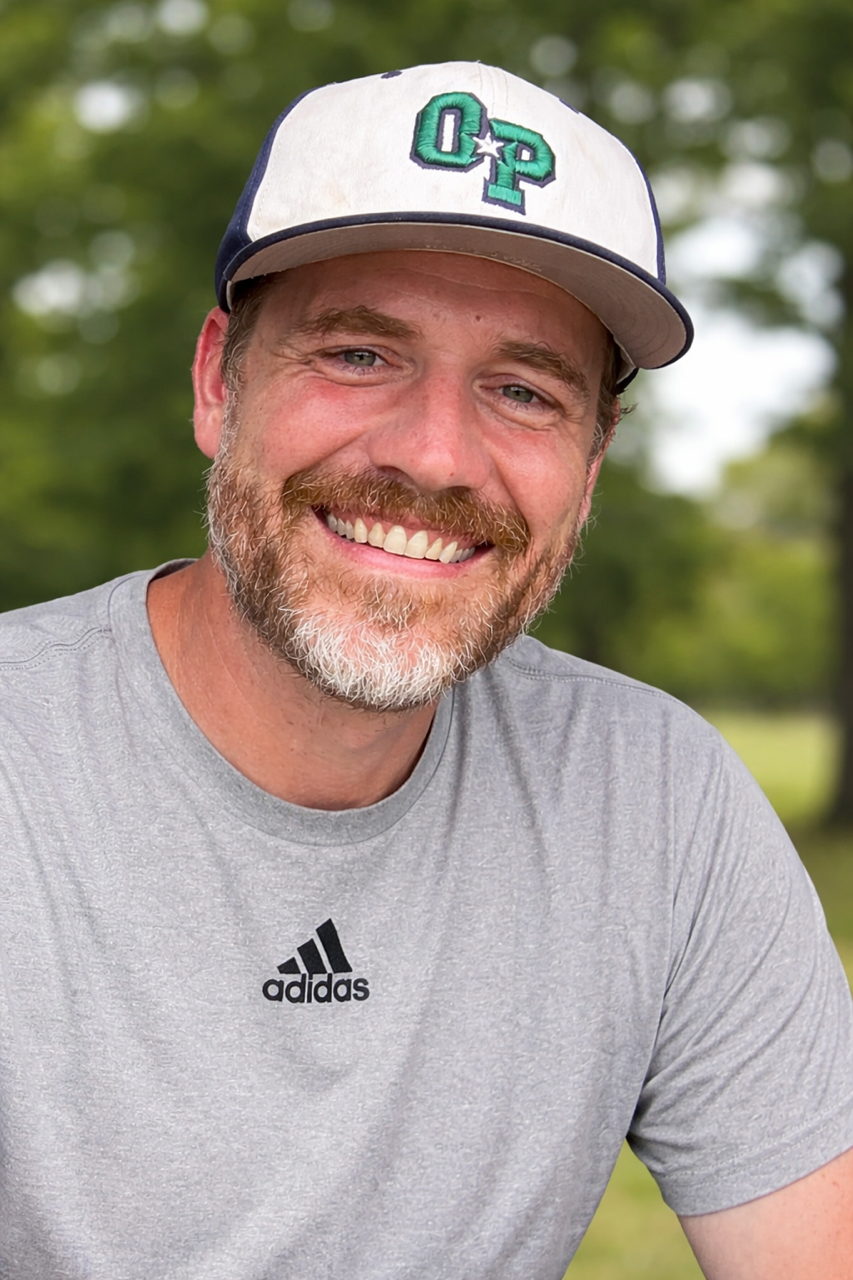 Football coach with arms crossed, looking stern on a sunny field, players in the background.