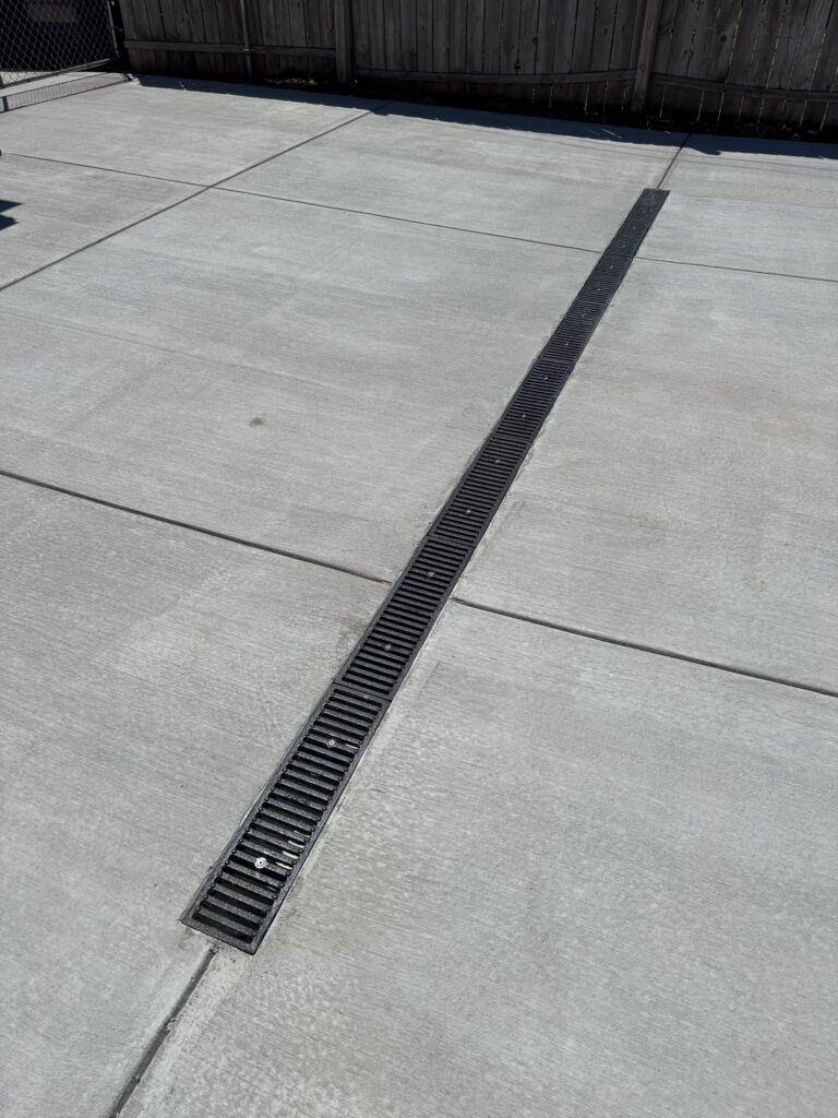 A drain on the side of a concrete driveway with a fence in the background.