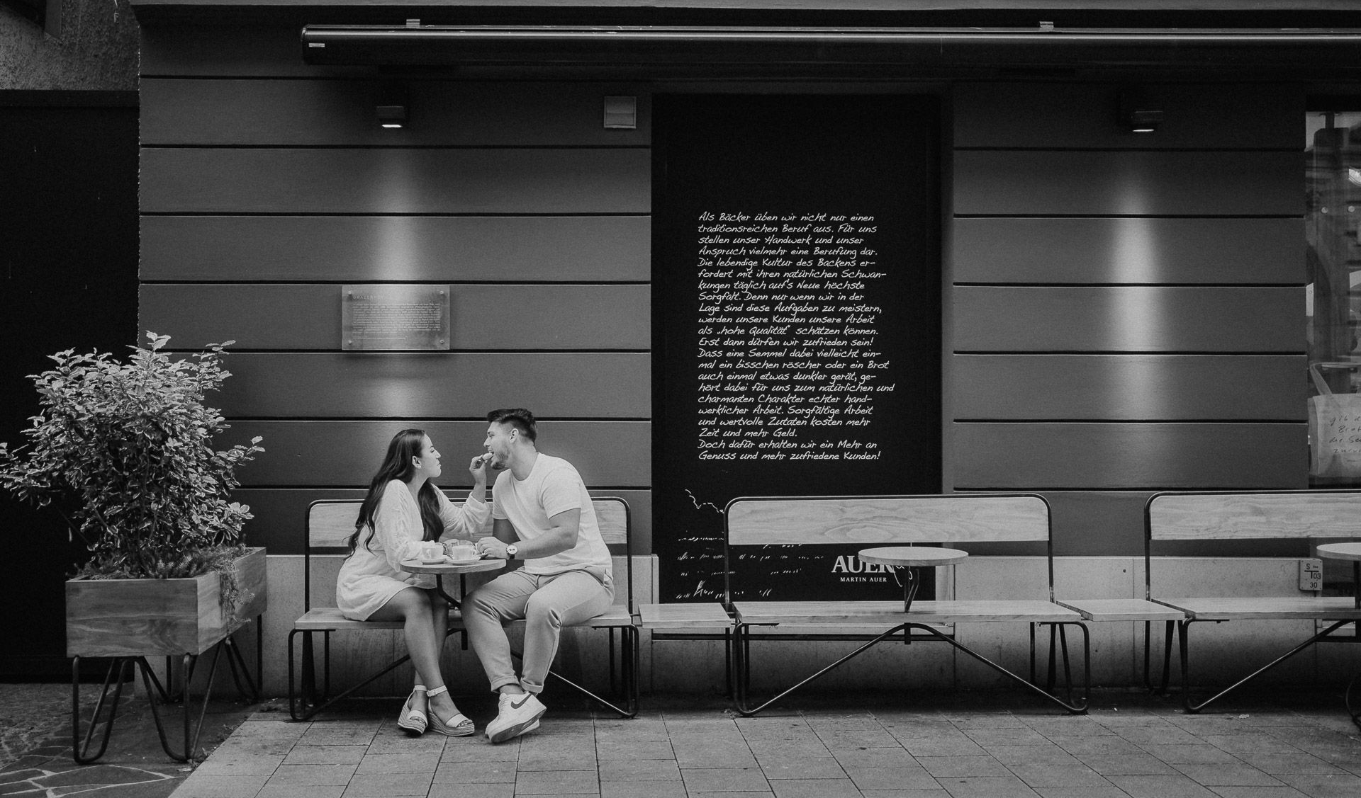 Couple sharing food while seated on a bench outside a building with text on the wall.