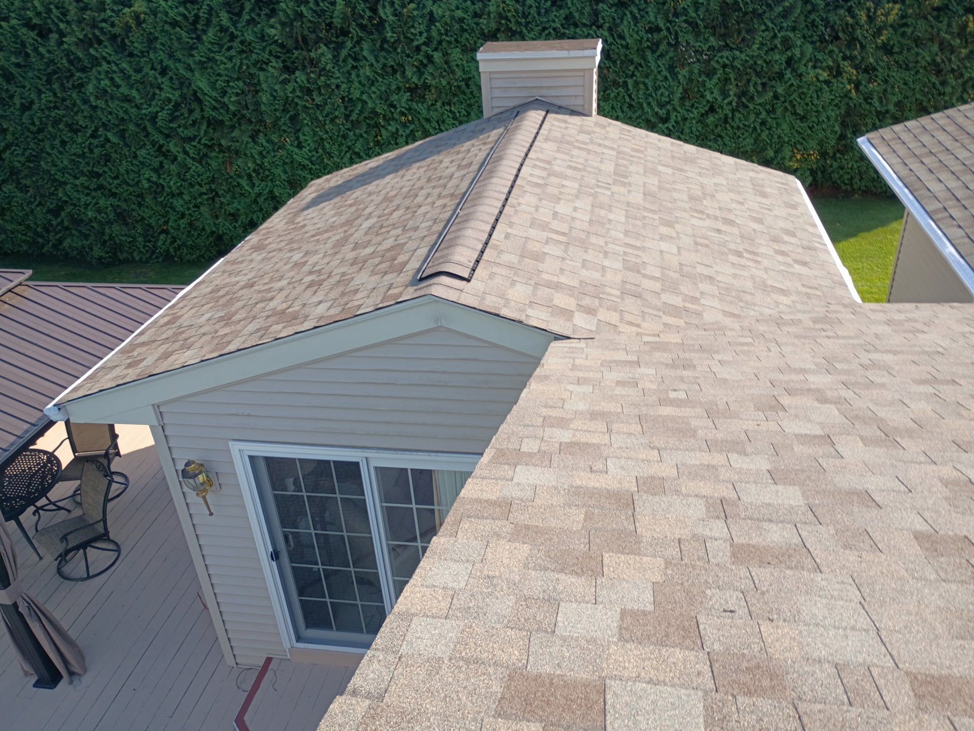 Tan roof with chimney against green bushes, adjacent to a deck with sliding glass door.
