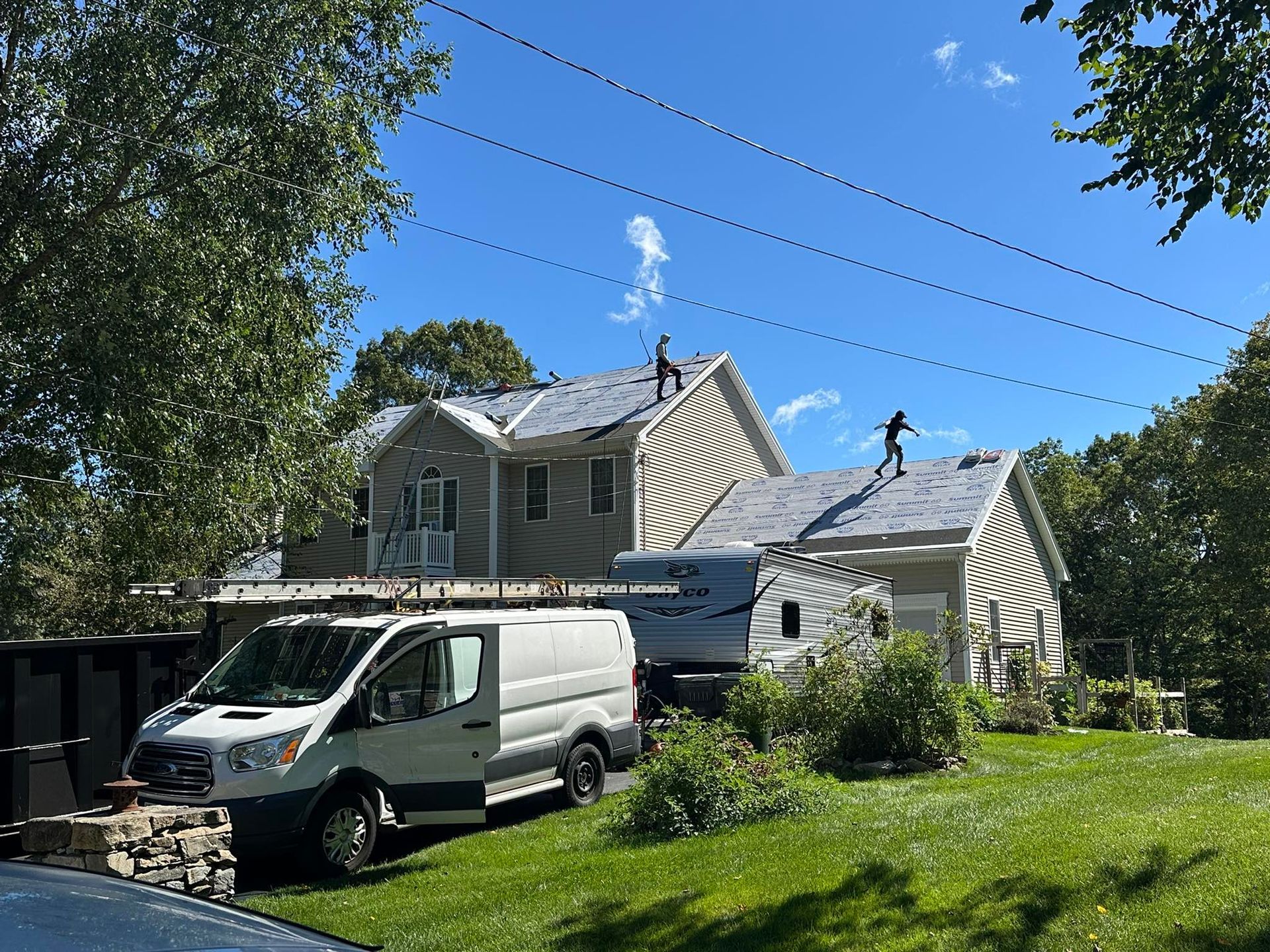 Roofers on a house, working on new shingles. A white van parked in front, sunny day.